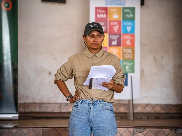 A woman in a cap holds a stack of papers as she presents, standing in front of a UN Sustainable Development Goals poster