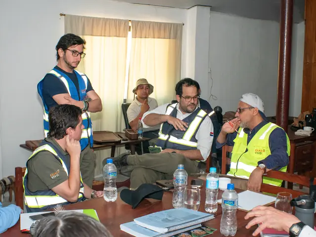 Team members in high-visibility vests sit and stand around a meeting table set with water bottles, folders and notebooks