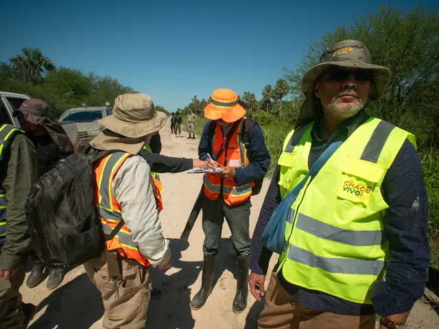 On a sandy track, a group in high-visibility vests and hats gathers around a person writing on a clipboard, with a parked vehicle behind