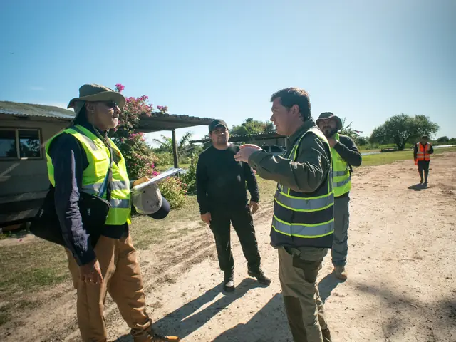 Team members in high-visibility vests talk beside a parked vehicle on a sandy road under a clear sky, one holding papers
