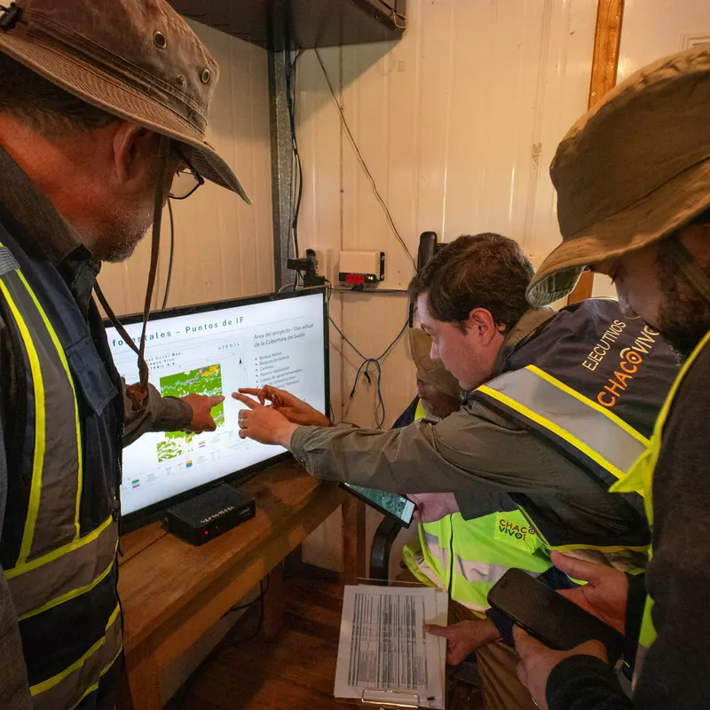 Team members in high-visibility vests lean in to a wall screen showing a project-area land-cover map, one pointing at the legend while others compare satellite imagery on their phones and hold a printed data sheet
